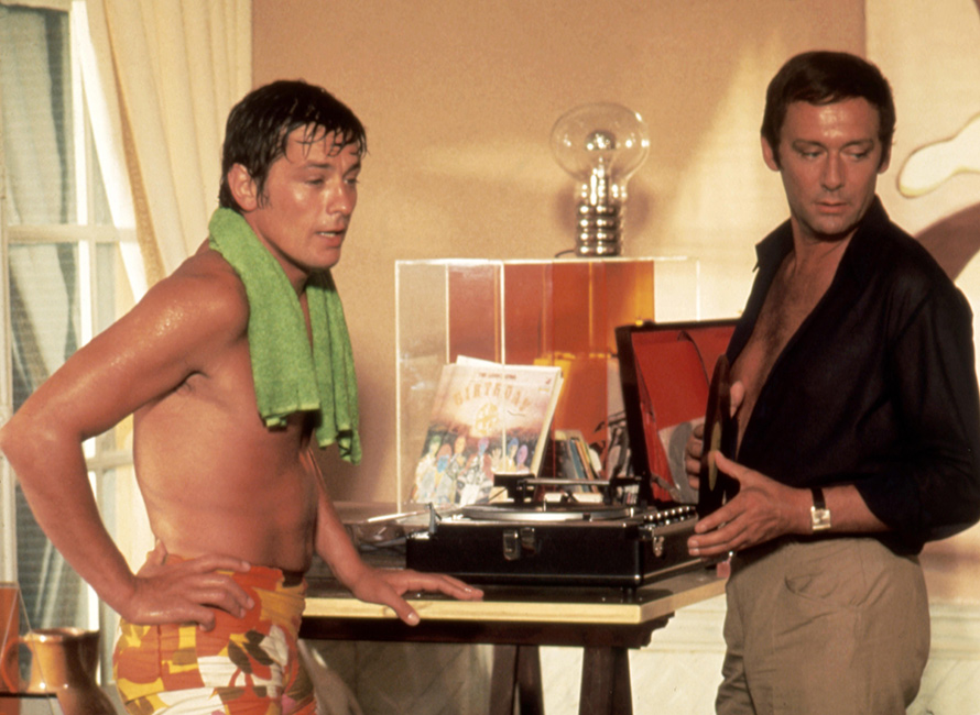 Alain Delon in costume fantasia e asciugamano verde accanto a un uomo in camicia nera