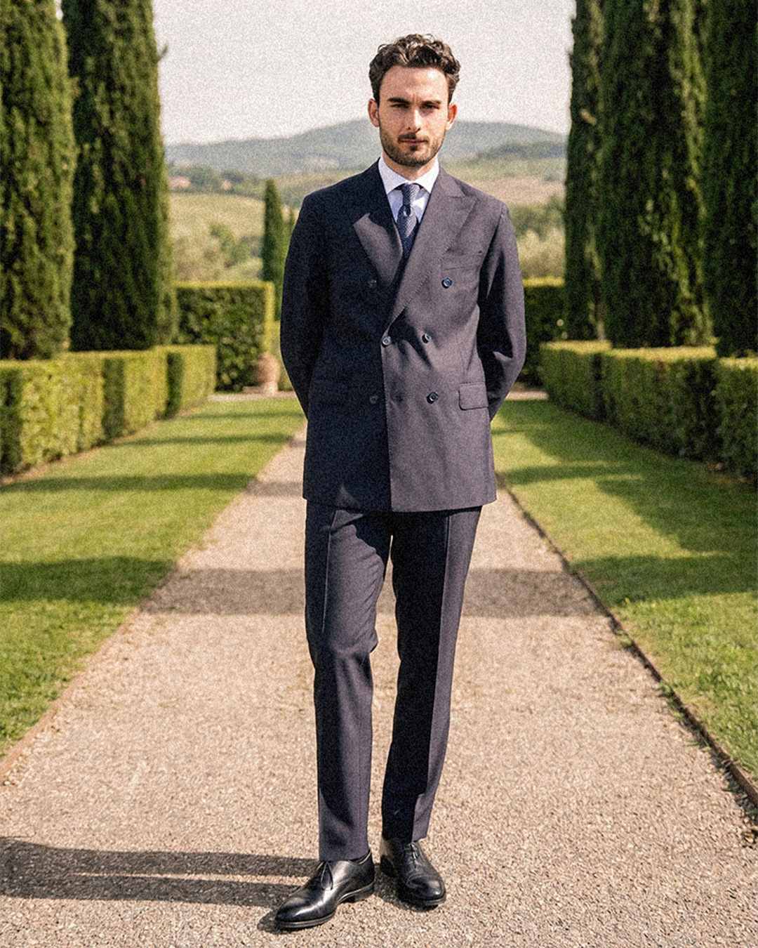 Uomo in abito doppiopetto blu scuro, camicia bianca e cravatta su un vialetto tra i cipressi