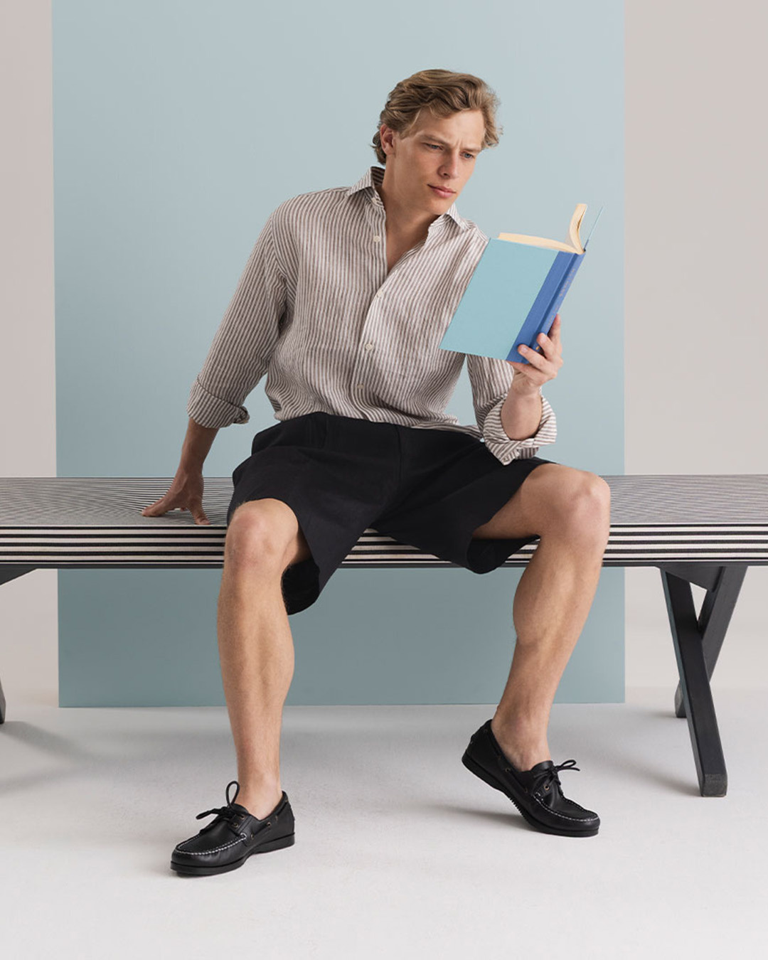 Uomo in camicia a righe e shorts neri seduto su una panca mentre legge un libro
