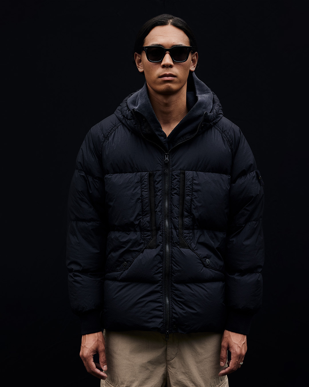 Uomo con occhiali da sole, piumino nero con cappuccio e pantaloni beige su sfondo nero