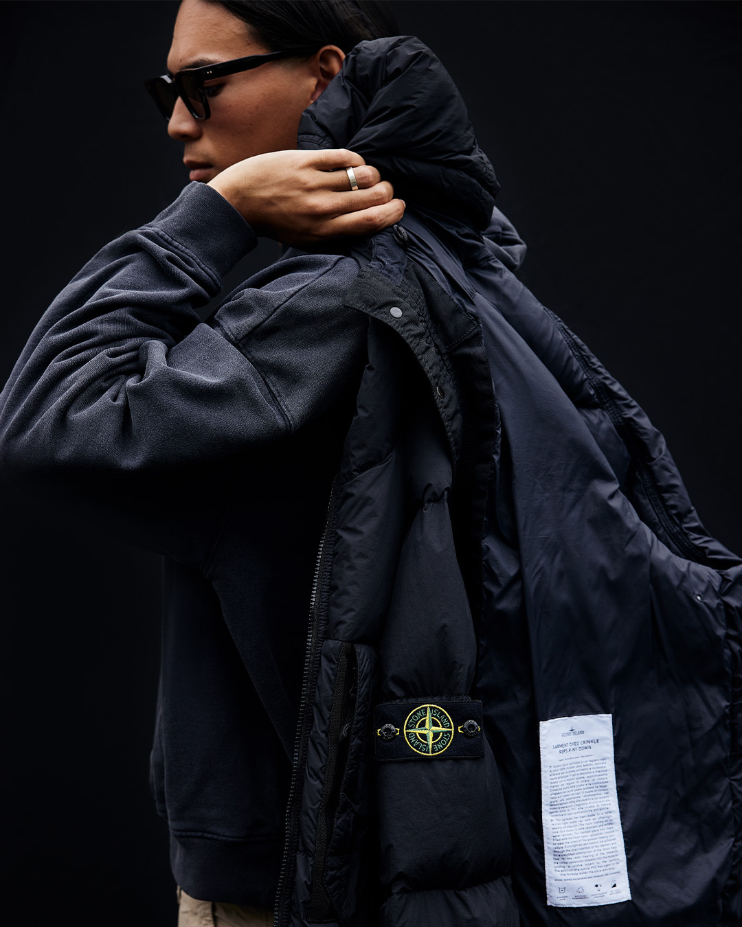 Uomo con occhiali da sole e giacca Stone Island scura con il badge della rosa dei venti sulla manica su sfondo nero