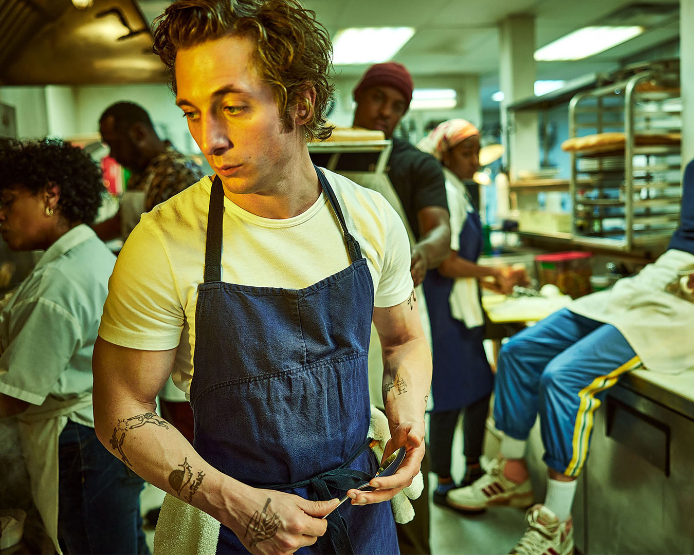 Il personaggio Carmy nella serie TV The Bear in t-shirt bianca e grembiule blu in una cucina di ristorante