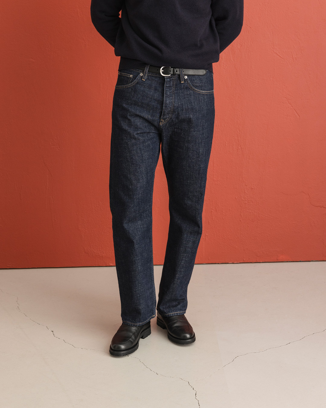 Uomo in jeans selvedge scuri dritti, maglione nero e mocassini neri davanti a una parete rossa