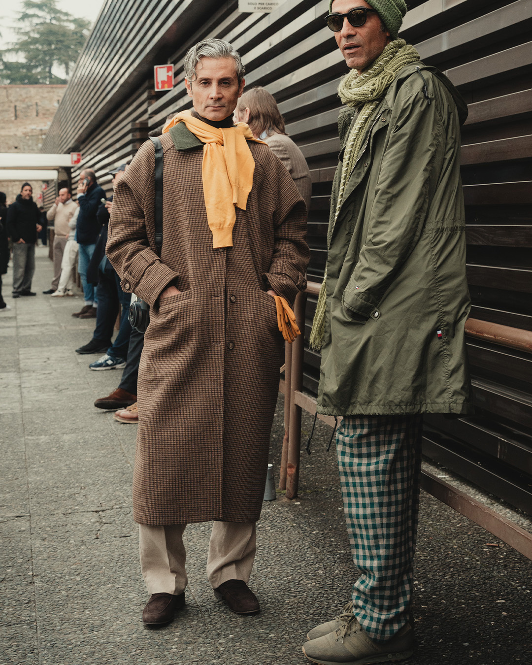 Due uomini in cappotto marrone e parka verde, uno con sciarpa arancione, in strada durante il Pitti Uomo