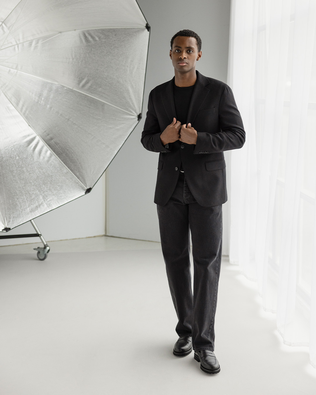 Uomo in giacca nera, maglione nero, pantaloni neri e mocassini in uno studio fotografico