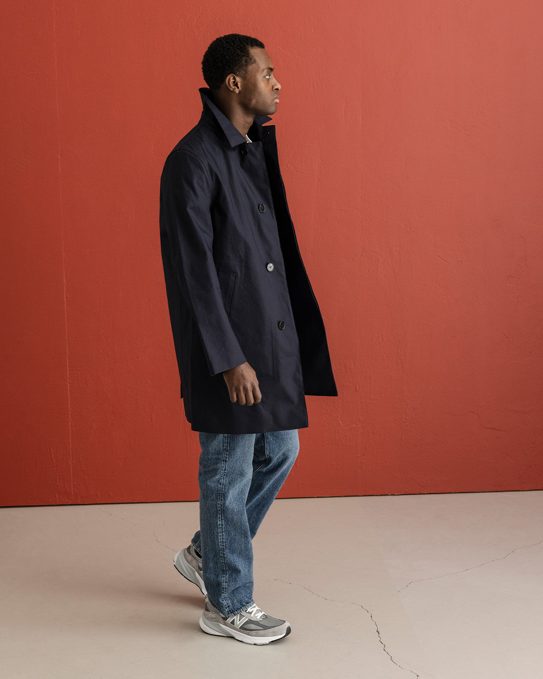 Uomo in cappotto blu scuro, jeans chiari e sneakers grigie davanti a una parete rossa
