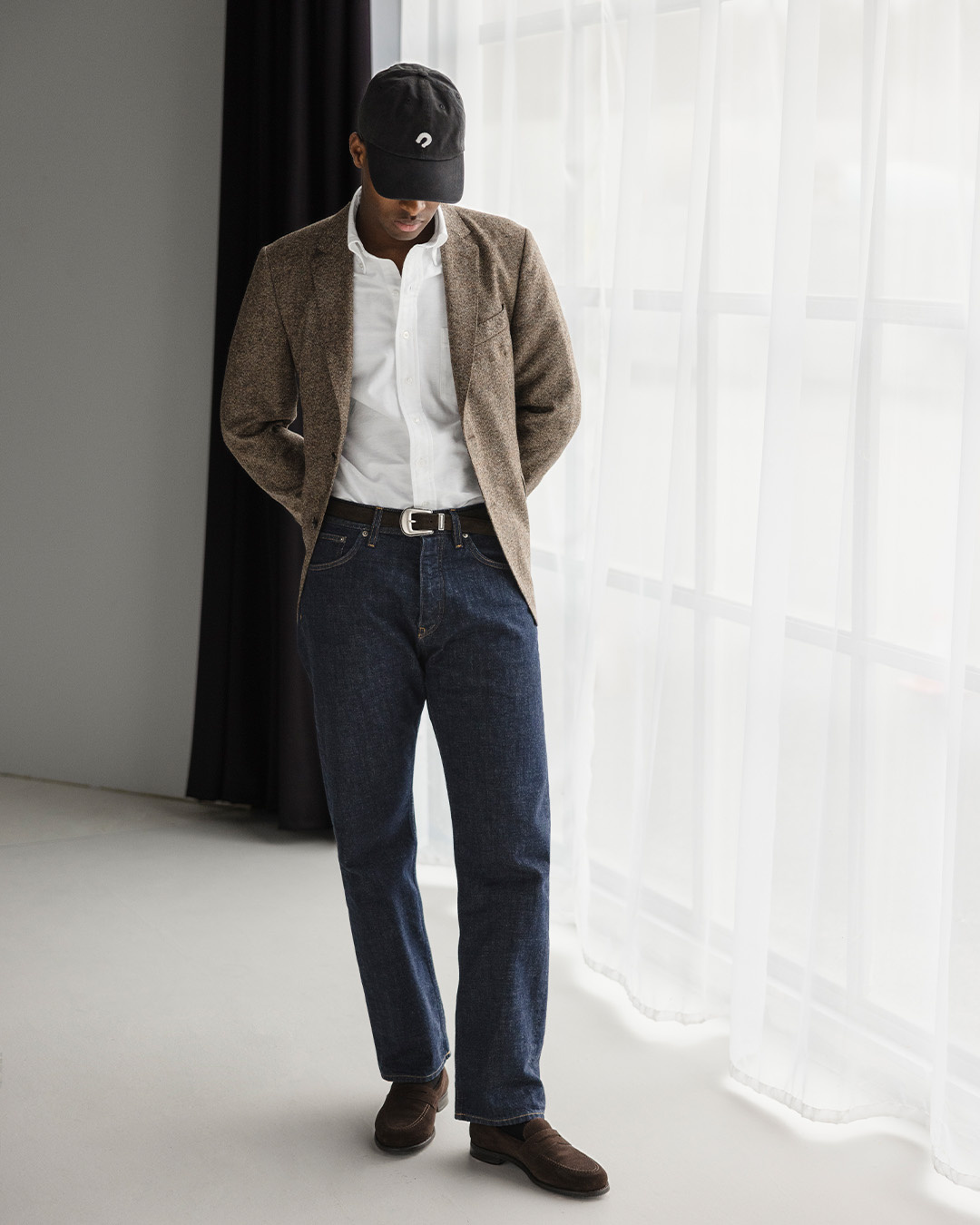 Uomo in giacca di tweed marrone, camicia bianca, cappellino, jeans in raw denim e mocassini marroni