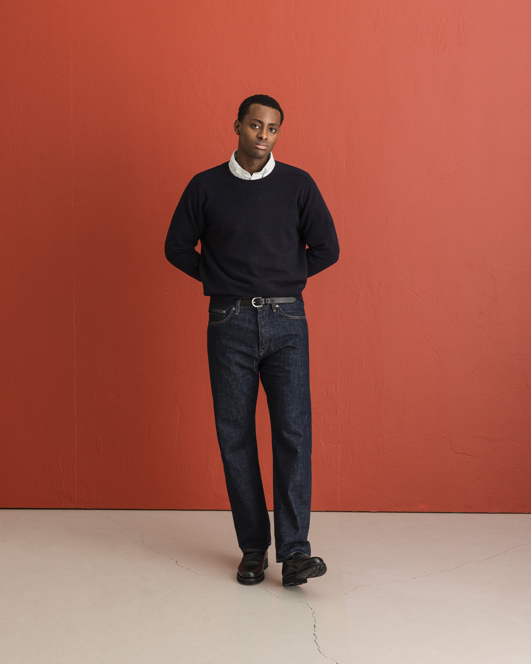 Uomo in pullover blu scuro, jeans in raw denim e mocassini neri davanti a una parete rossa