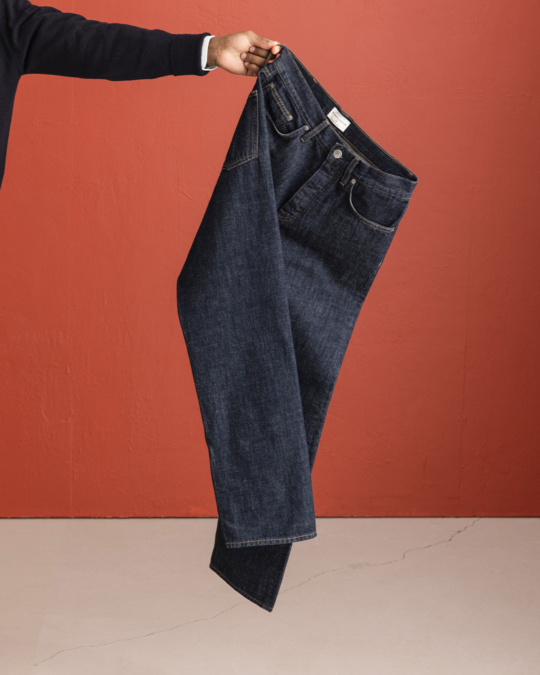Mano che regge un paio di jeans scuri in raw denim davanti a una parete rossa