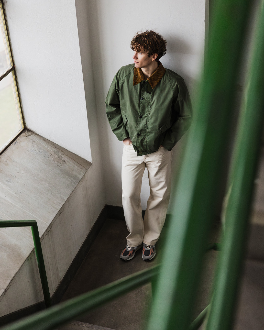 Uomo in giacca cerata verde e pantaloni chiari appoggiato a una parete in una tromba delle scale