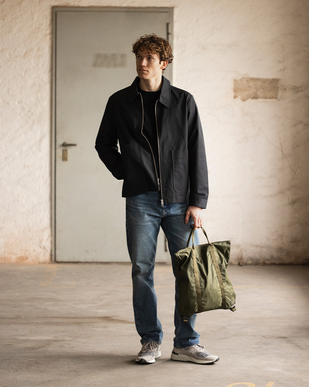 Uomo in giacca in nylon nera, jeans e sneakers con una borsa verde