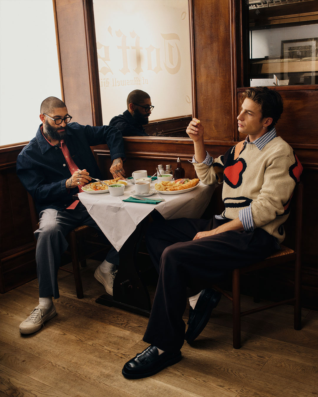 Due uomini a un tavolo di ristorante, uno in maglione chiaro con motivo a cuore e pantaloni scuri
