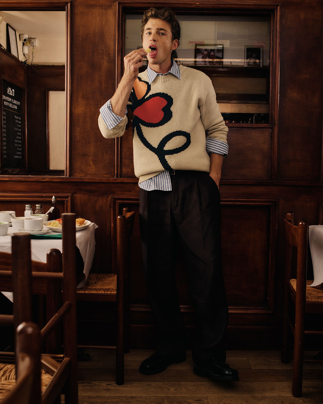 Uomo in maglione chiaro con cuore rosso, camicia a righe e pantaloni sartoriali scuri in un ristorante