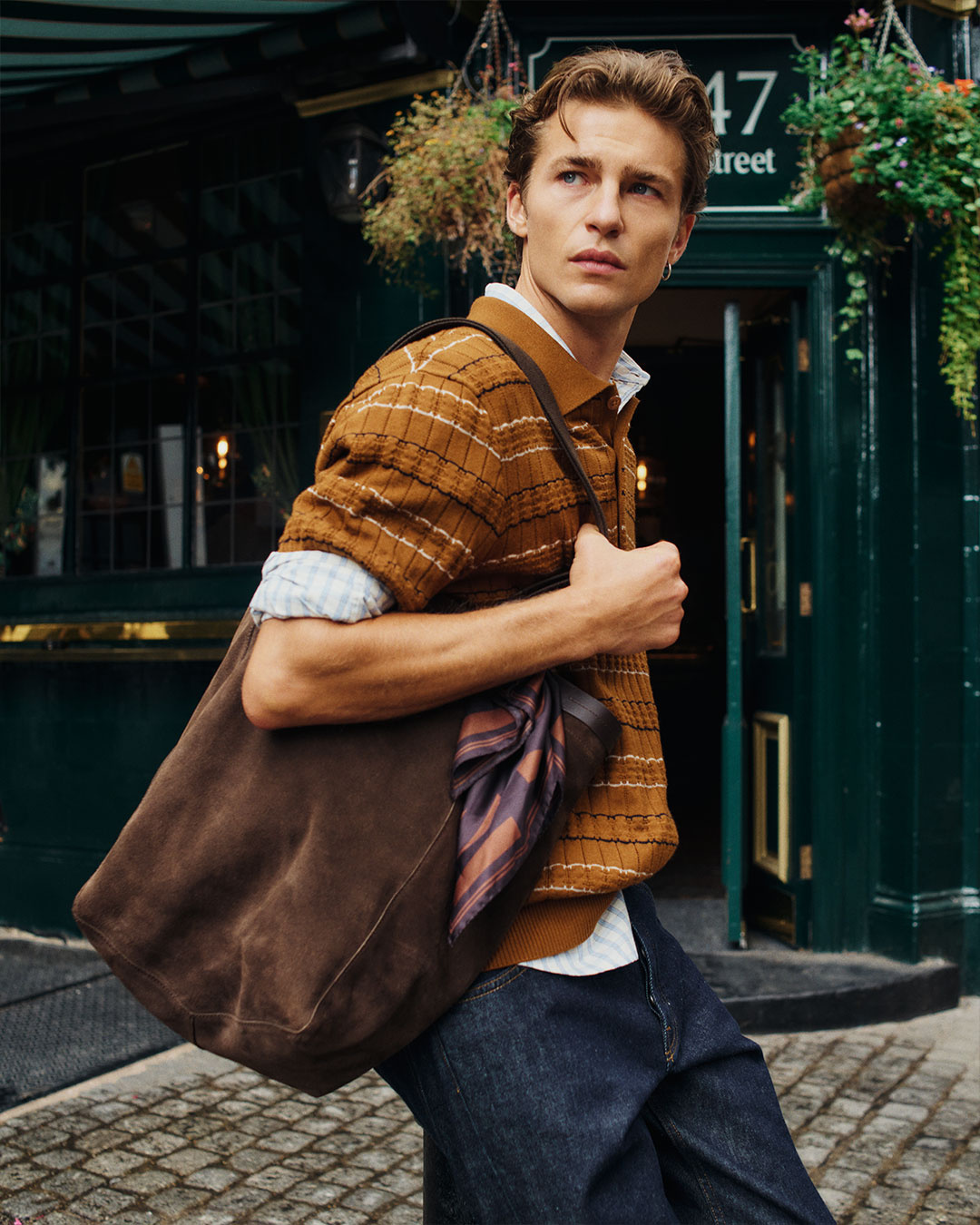 Uomo in polo in maglia a righe marroni e jeans scuri con una borsa marrone in spalla
