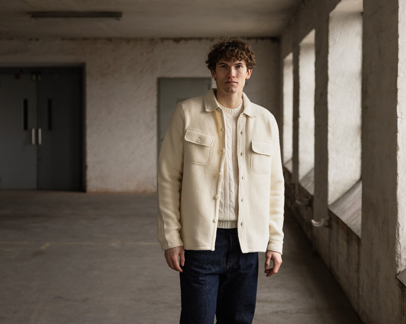 Uomo in overshirt color crema e jeans scuri in un corridoio luminoso