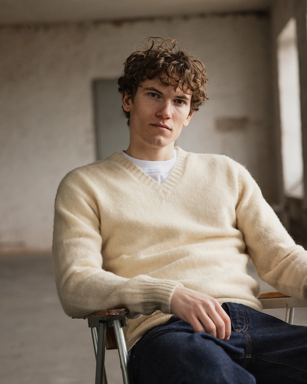 Uomo in maglione color crema con scollo a V in lana d'agnello e jeans scuri seduto su una sedia