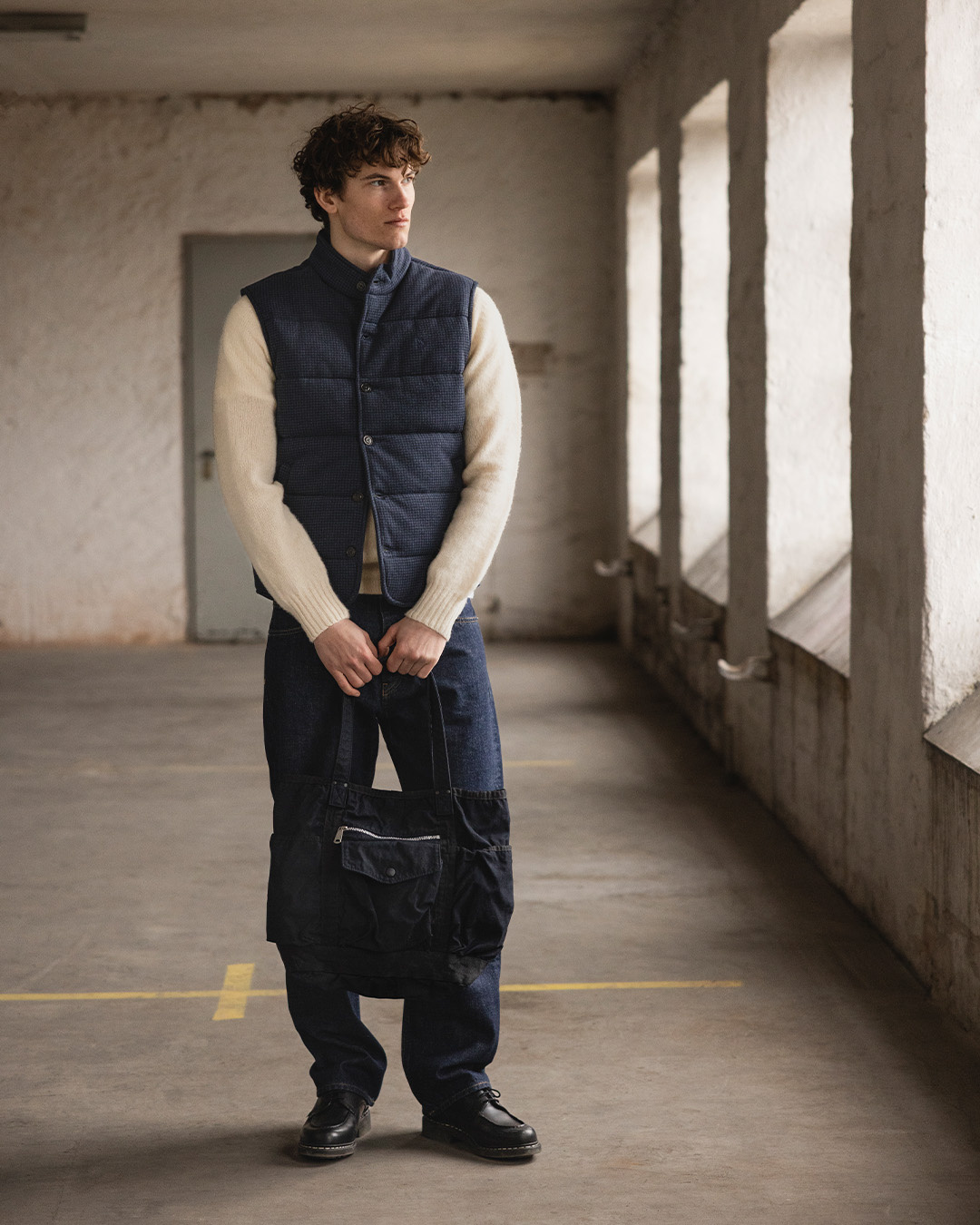Uomo in gilet trapuntato, maglione chiaro, jeans e derby neri con una borsa