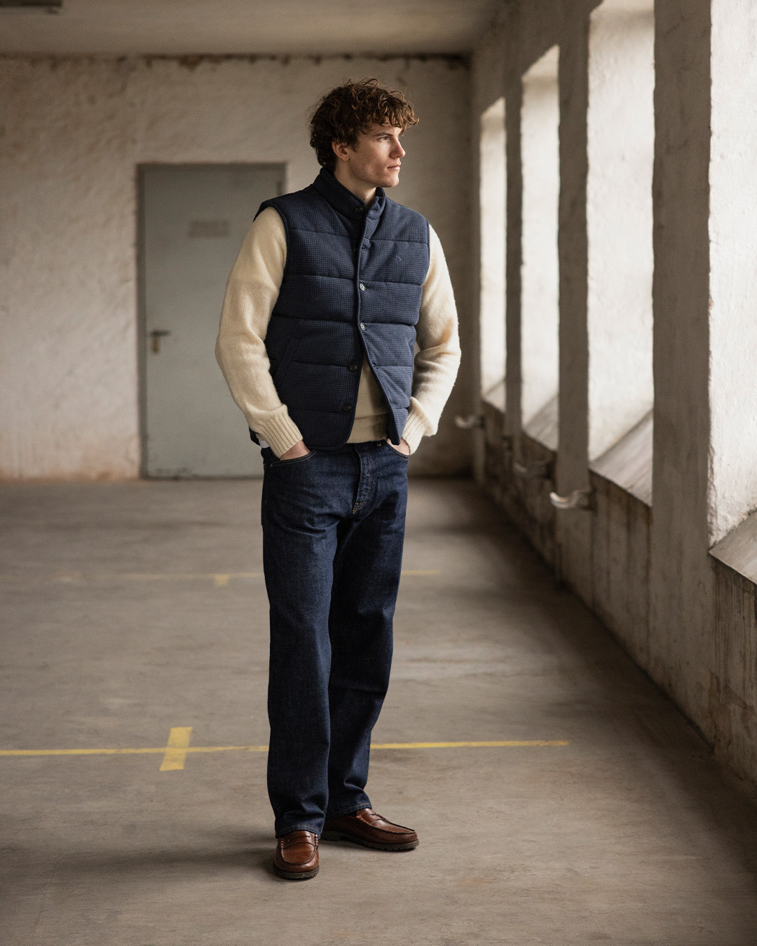 Uomo in gilet trapuntato blu navy, maglione chiaro, jeans e mocassini marroni