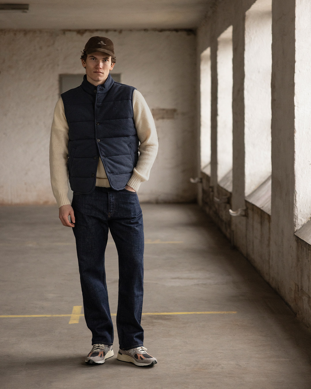 Uomo con cappellino, gilet trapuntato blu navy, maglione chiaro, jeans e sneakers