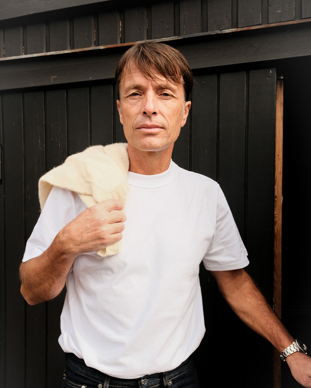 Uomo in t-shirt bianca con un maglione sulla spalla davanti a una parete di legno nera