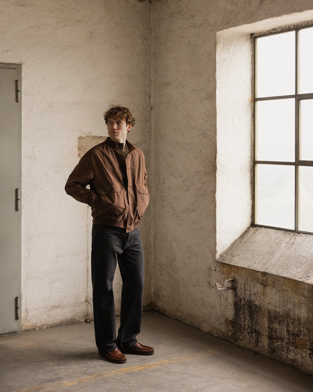 Uomo in giacca marrone, maglione bianco crema, pantaloni neri e mocassini marroni in una stanza luminosa