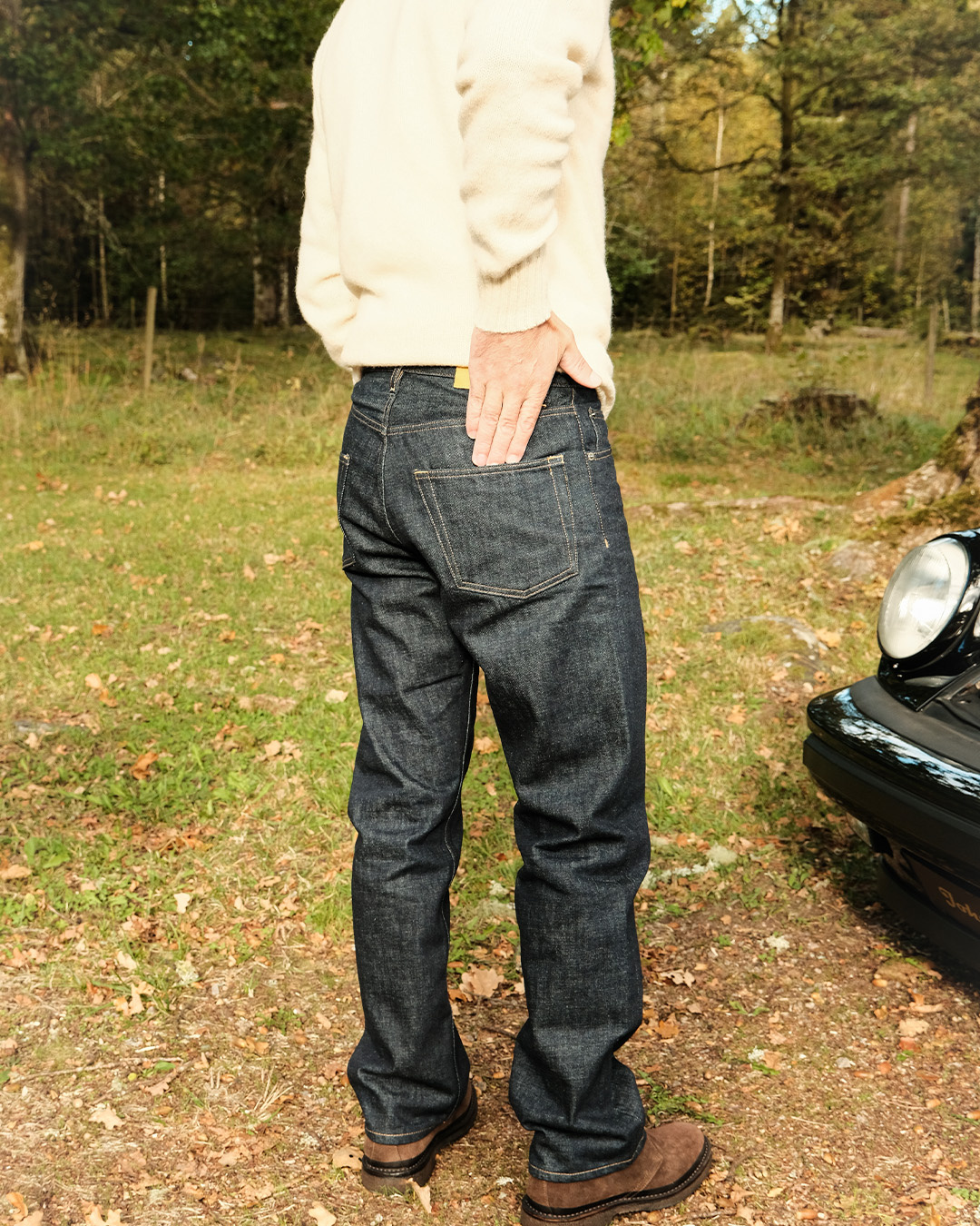Uomo in jeans scuri e maglione chiaro visto di spalle in un prato accanto a un'auto