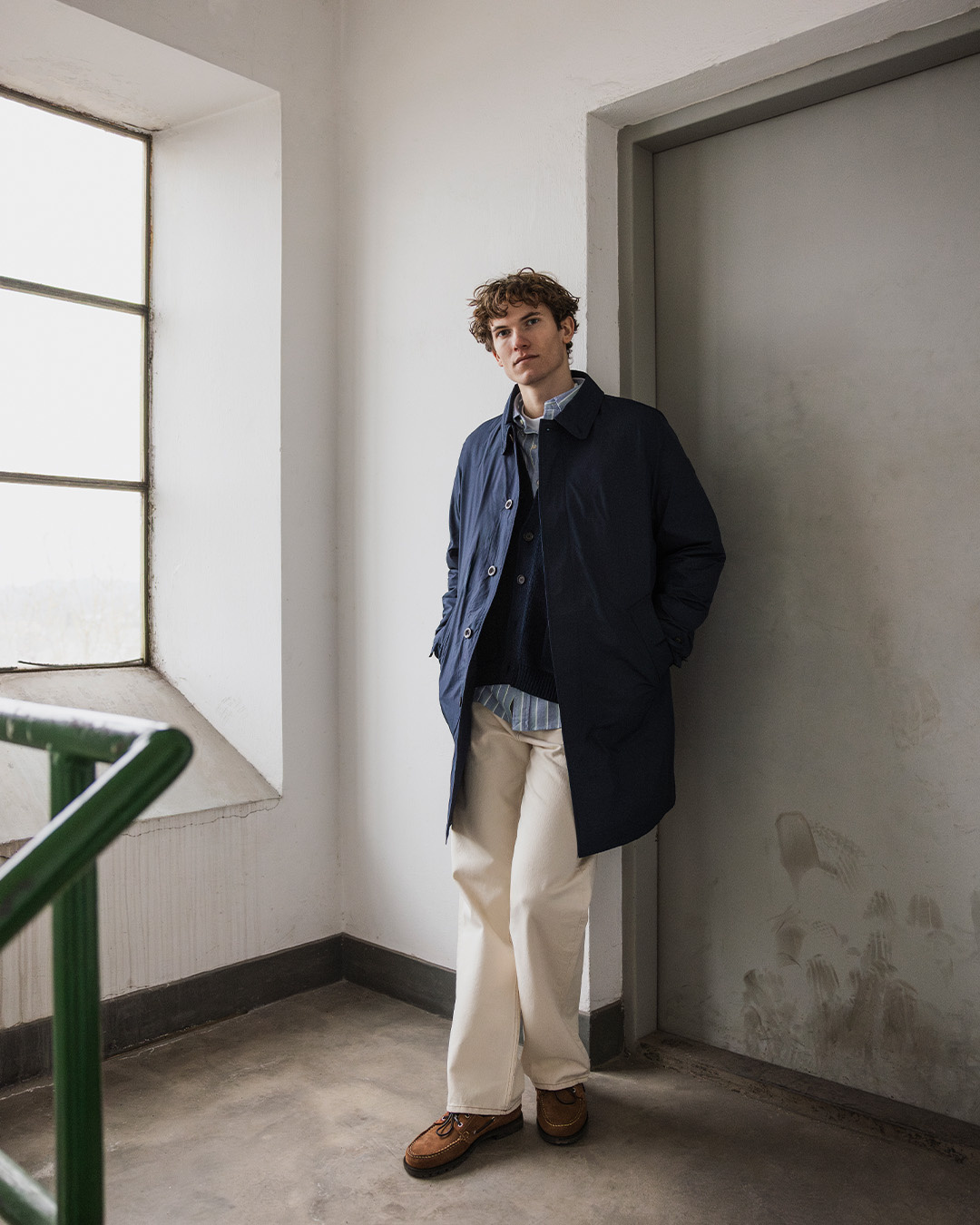 Uomo in windbreaker lungo blu navy e pantaloni chiari accanto a una parete