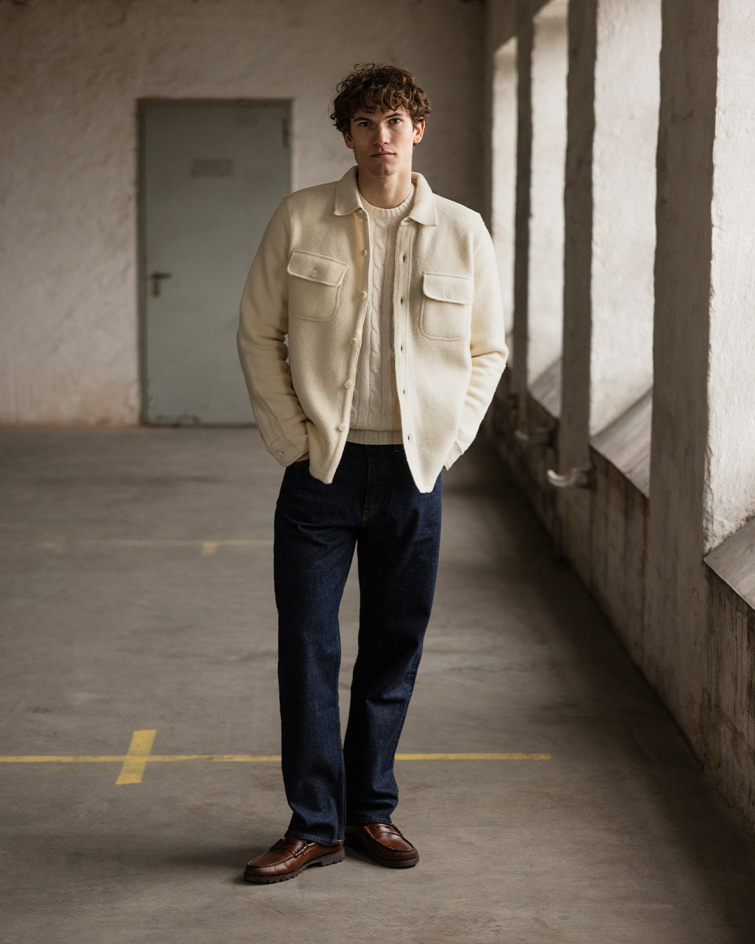 Uomo in overshirt color crema, pantaloni scuri e scarpe marroni in un corridoio