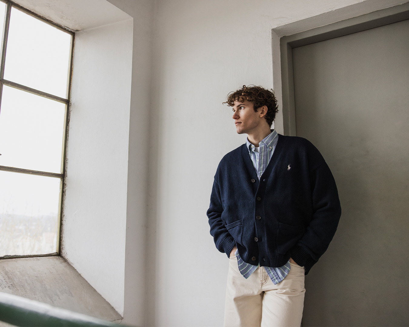 Uomo in cardigan blu navy e pantaloni chiari appoggiato a una parete