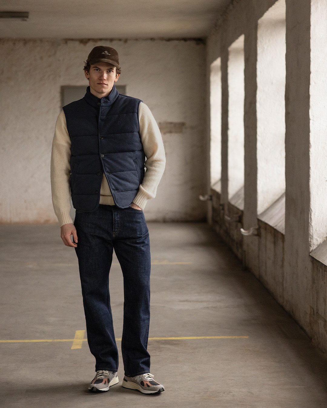 Uomo con cappellino, gilet blu navy e pantaloni scuri in un corridoio