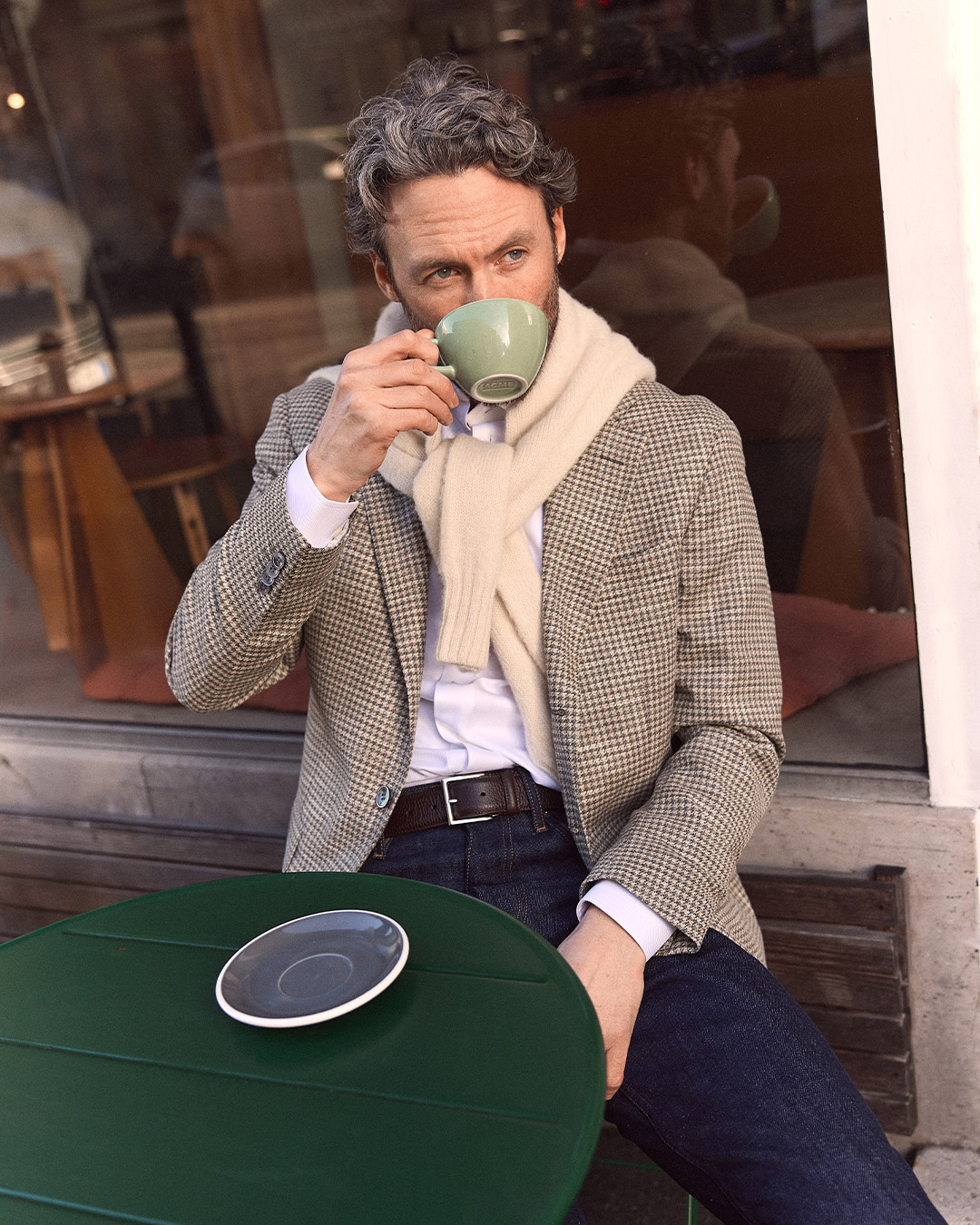 Uomo in giacca a quadri con un maglione sulle spalle che beve un caffè a un tavolo
