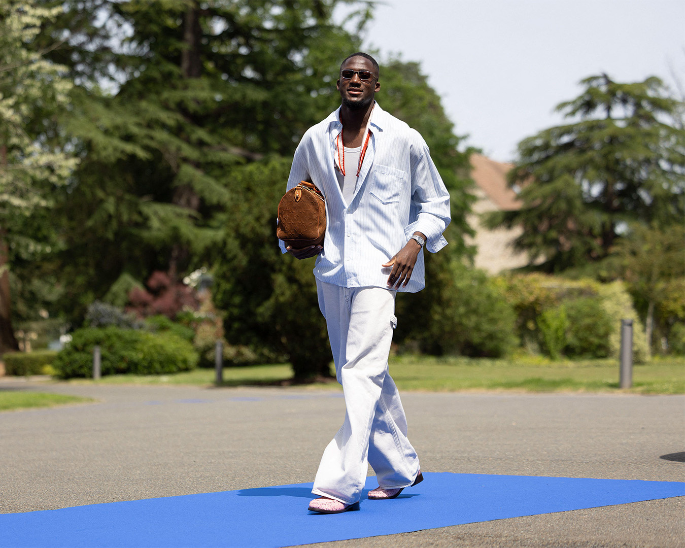 Ibrahima Konaté vestito di chiaro con una borsa Louis Vuitton sotto il braccio