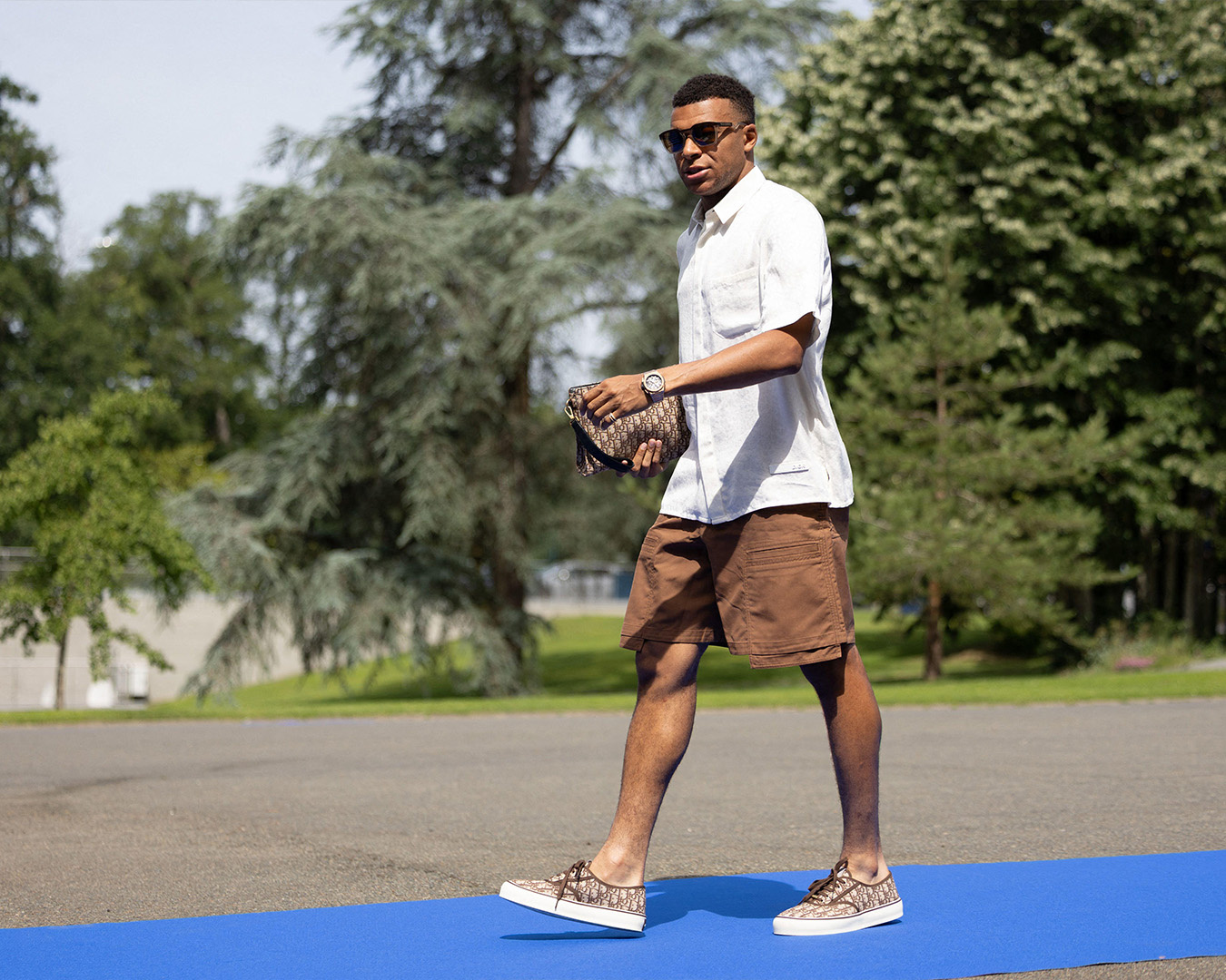 Kylian Mbappé in camicia bianca e shorts marroni con una clutch Dior