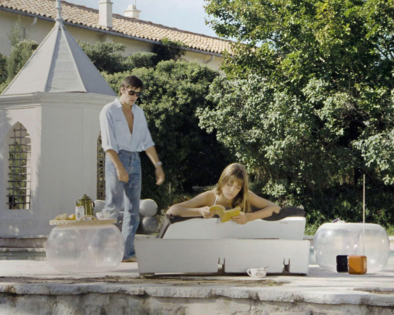 Alain Delon in camicia chiara a bordo piscina con una donna su un lettino in La Piscine