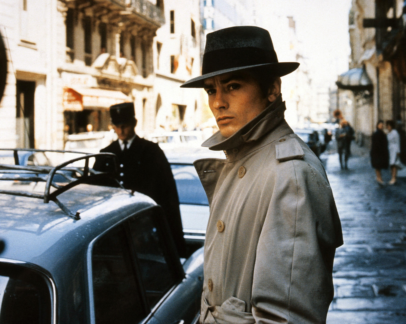 Alain Delon in trench chiaro e fedora in una strada in Le Samouraï