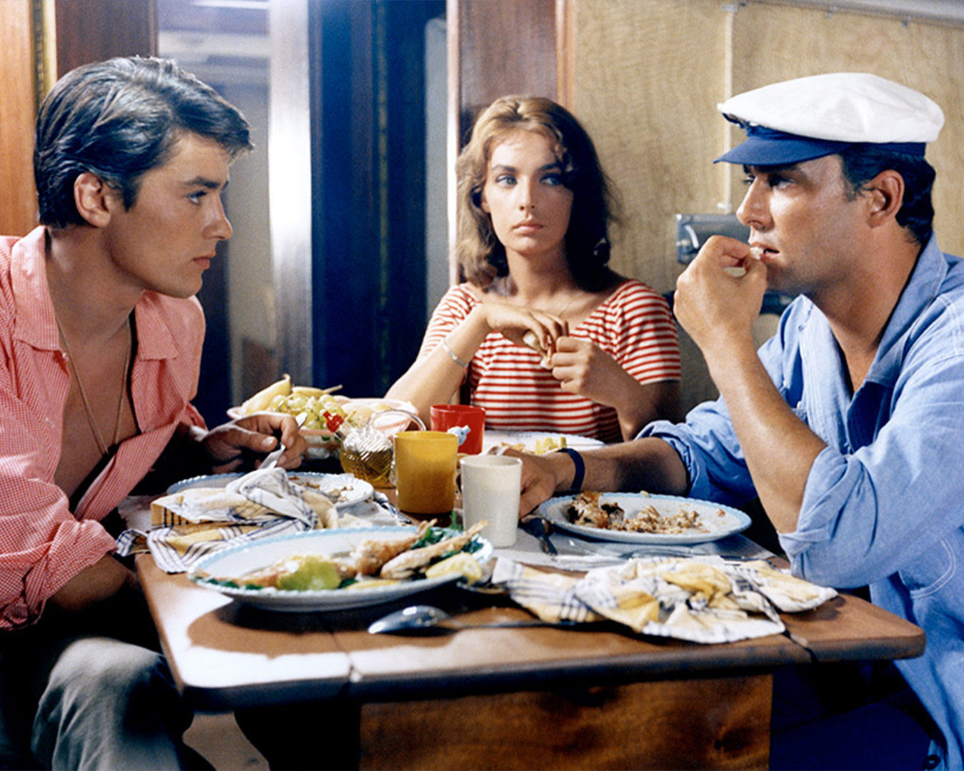 Alain Delon in camicia rosa a un tavolo da pranzo con una donna e un uomo con berretto in Plein Soleil