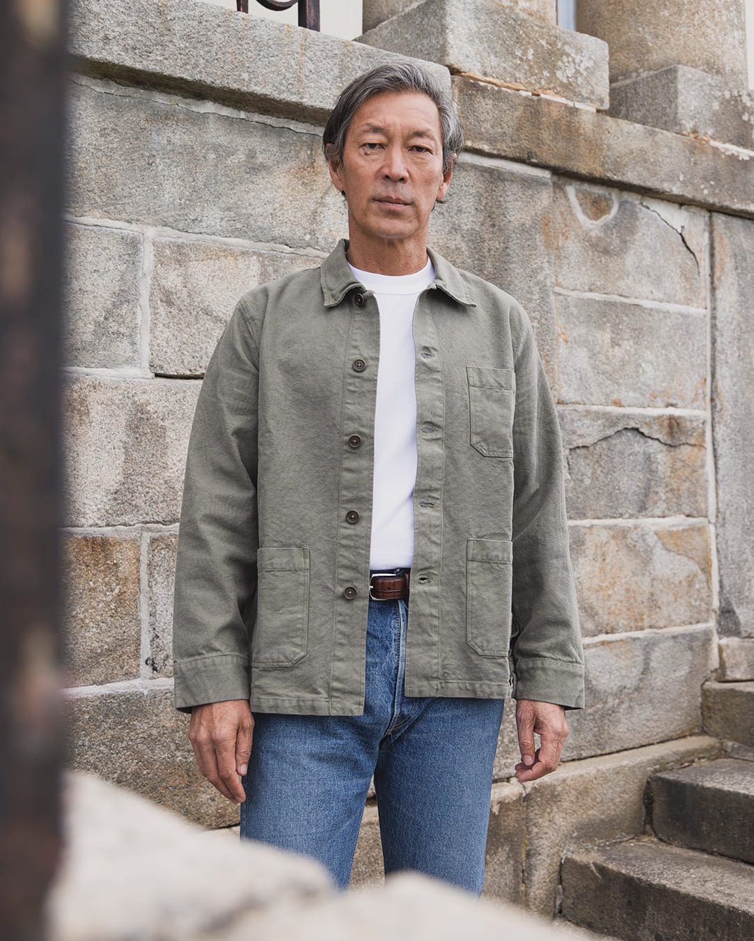 Uomo in overshirt verde, t-shirt bianca e jeans davanti a un muro di pietra