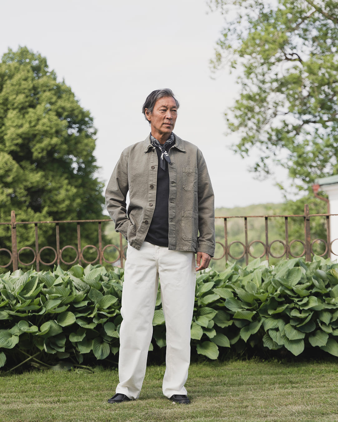 Uomo in overshirt verde, foulard e jeans bianchi in un giardino