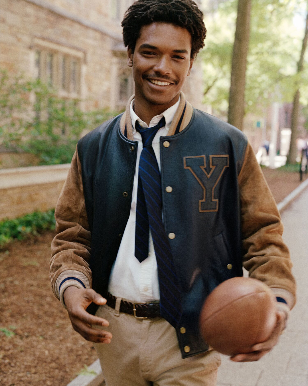 Ragazzo sorridente in giacca varsity con la Y e cravatta che tiene un pallone da football americano