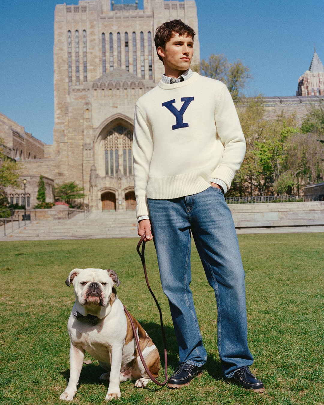 Uomo in maglione bianco con la Y e jeans con un bulldog inglese davanti a un edificio di Yale