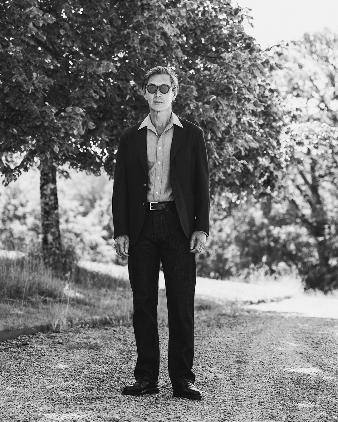 Foto in bianco e nero di un uomo in giacca, camicia e occhiali da sole su un sentiero di ghiaia