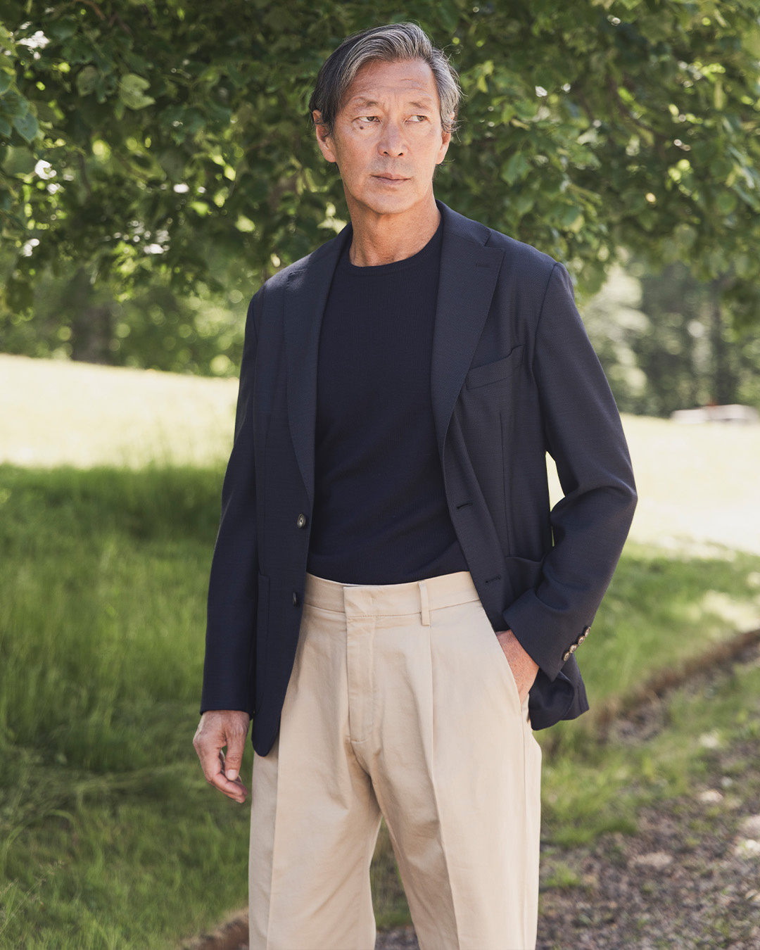 Uomo in giacca blu scuro, t-shirt nera e pantaloni beige in un giardino