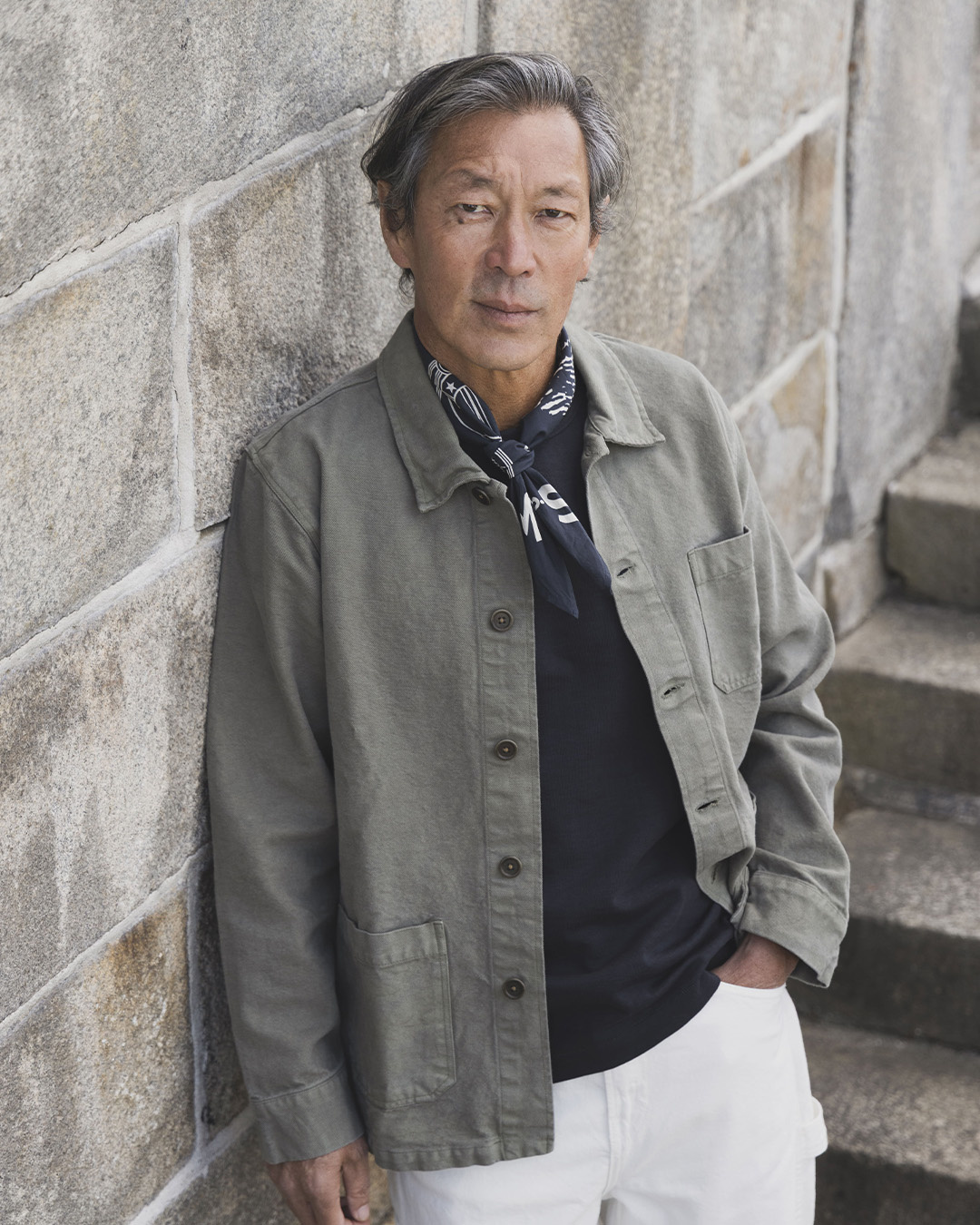Uomo in overshirt verde e foulard al collo appoggiato a un muro di pietra