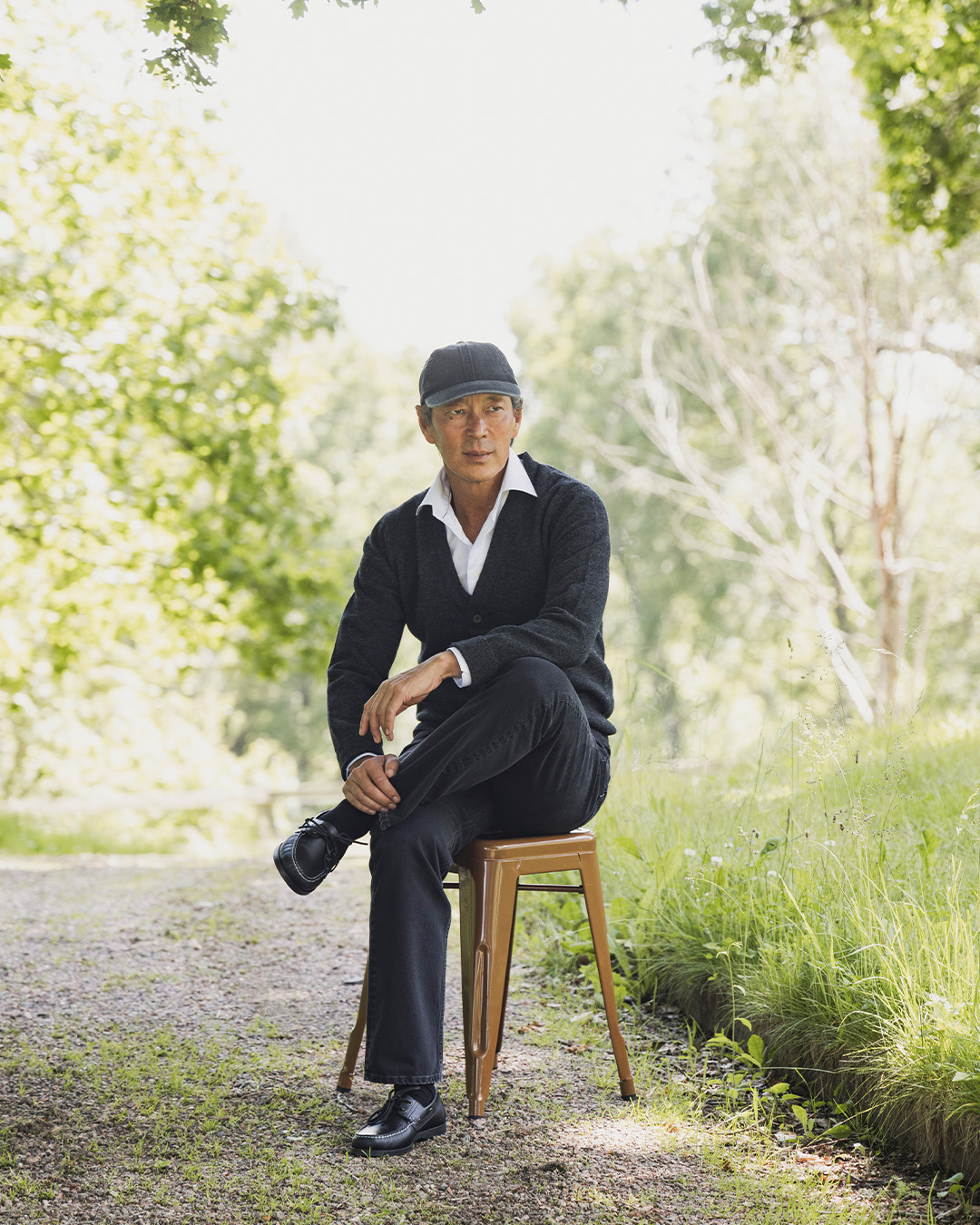 Uomo con cappellino, cardigan scuro e jeans seduto su uno sgabello su un sentiero