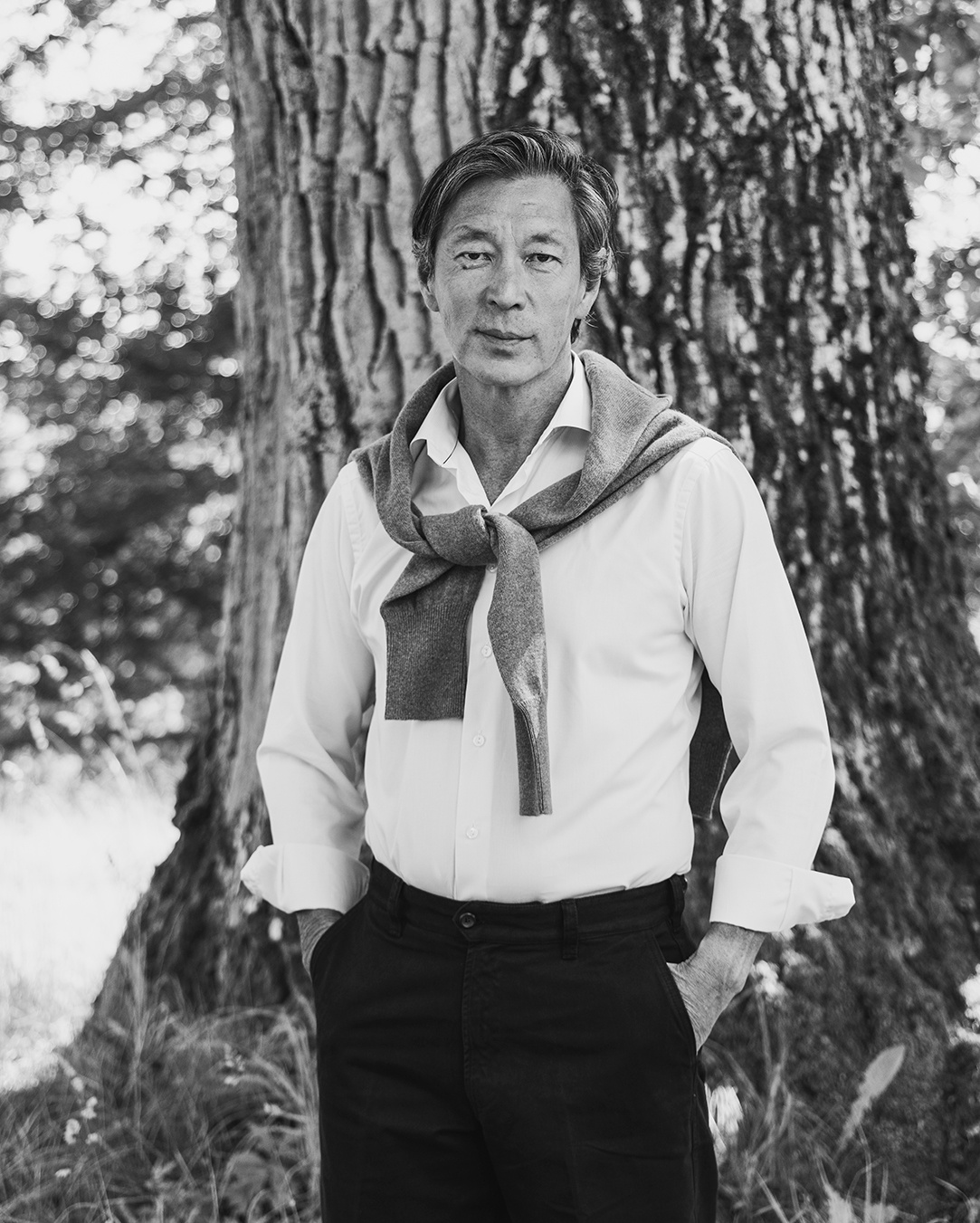 Foto in bianco e nero di un uomo in camicia con un maglione sulle spalle accanto a un albero