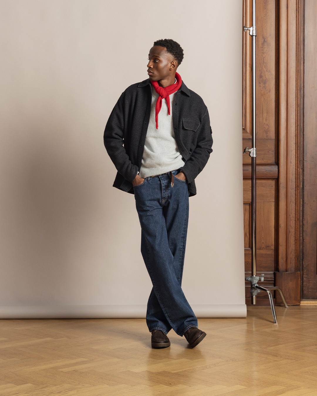 Uomo con overshirt scura, maglione grigio e sciarpa rossa, jeans dritti e chukka boot in camoscio marrone scuro